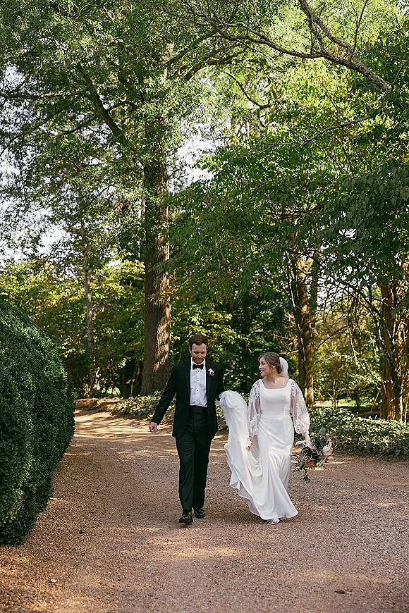 Couple portrait of bride and groom walking hand in hand, bride with veil and bouquet, along a sunlit tree-lined forest path