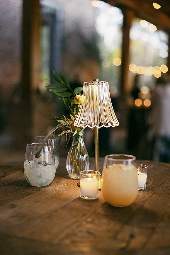 Wedding table decor with a wedding table lamp, votive candles, bud vase greenery and yellow flower on a wooden table against string lights and brick wall