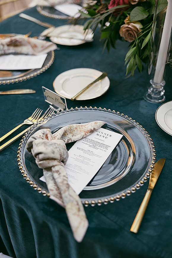 Reception tablescape with wedding place setting on dark tablecloth, beaded charger, gold flatware, menu card, taper candle, and floral greenery centerpiece