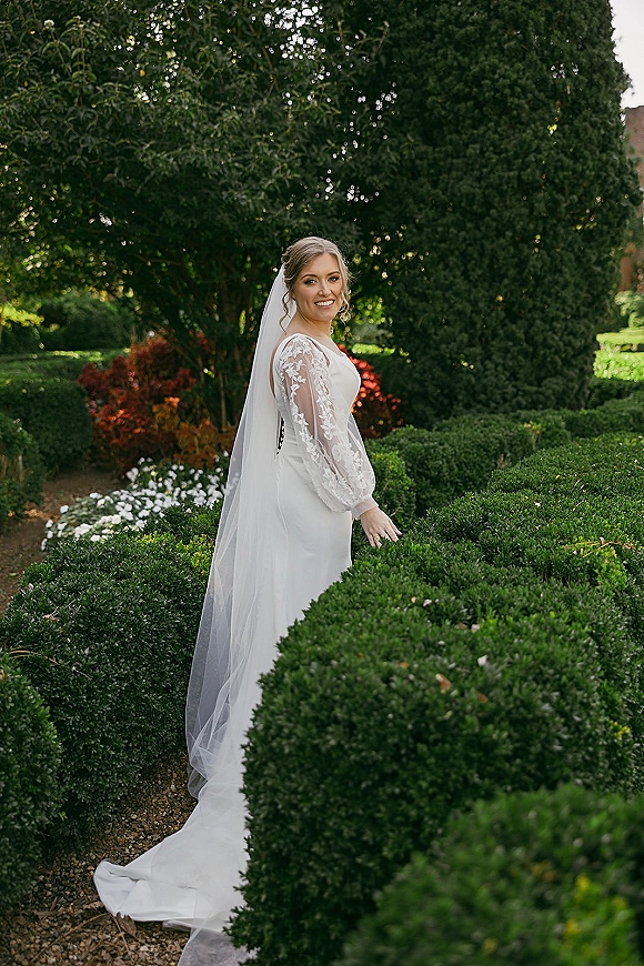 Bridal portrait of a bride looking back on a garden path, wearing a lace long sleeve wedding dress and cathedral veil by hedges