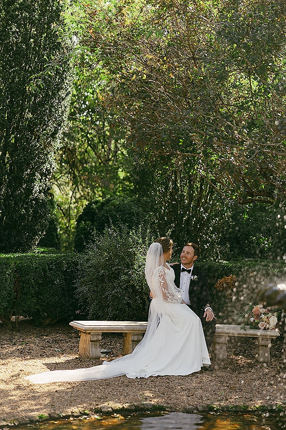 Couple portrait of bride sitting on groom lap on a stone bench, smiling in a garden by pond water, veil and peach bouquet visible