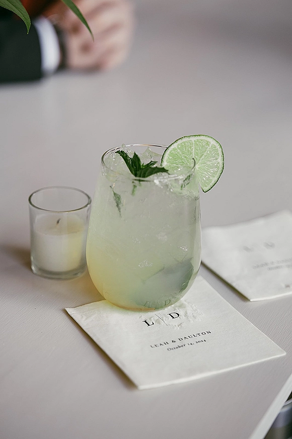Signature wedding cocktail beside a signature drink sign, served in a glass with ice, lime and mint on a reception table by guest hands