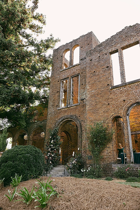 Ceremony entrance decor with a floral arch entrance of greenery and white-blush blooms, candles lining stone steps at brick ruins doorway