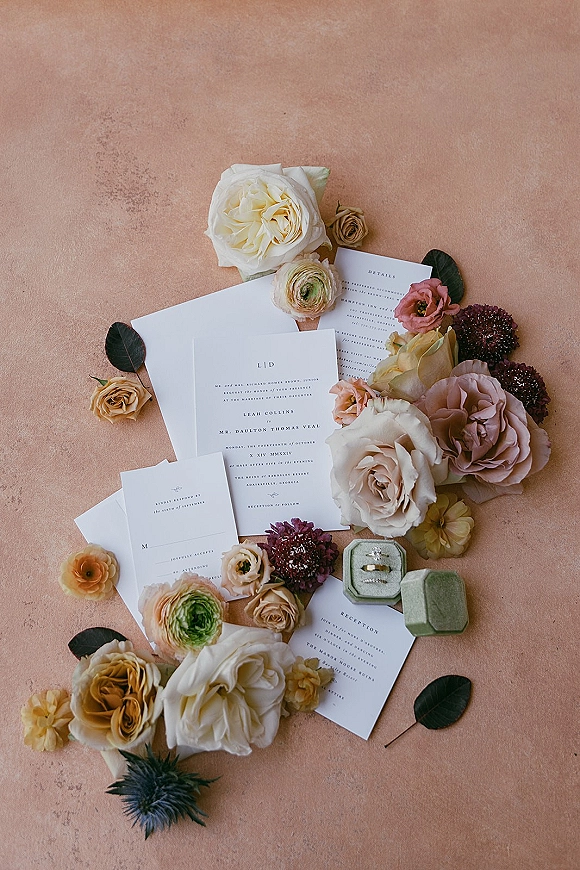 Invitation suite wedding invitation flat lay with envelopes, RSVP and reception cards, roses and ranunculus, plus ring box and rings on peach backdrop