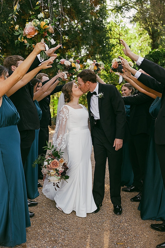 Wedding kiss portrait of newlyweds kissing under a tree canopy, framed by bridal party tunnel with bouquets, bride’s lace sleeves and veil