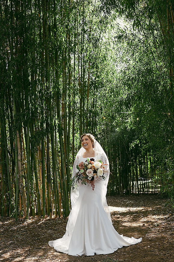 Bridal portrait of a bride holding bouquet, her long veil and white dress train flowing as she walks a sunlit bamboo forest path