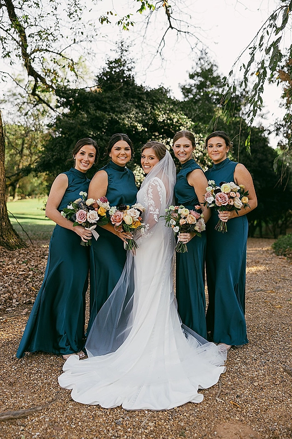 Bride and bridesmaids in teal dresses stand on a garden path, holding blush and mauve bouquets as the bride’s veil trails behind