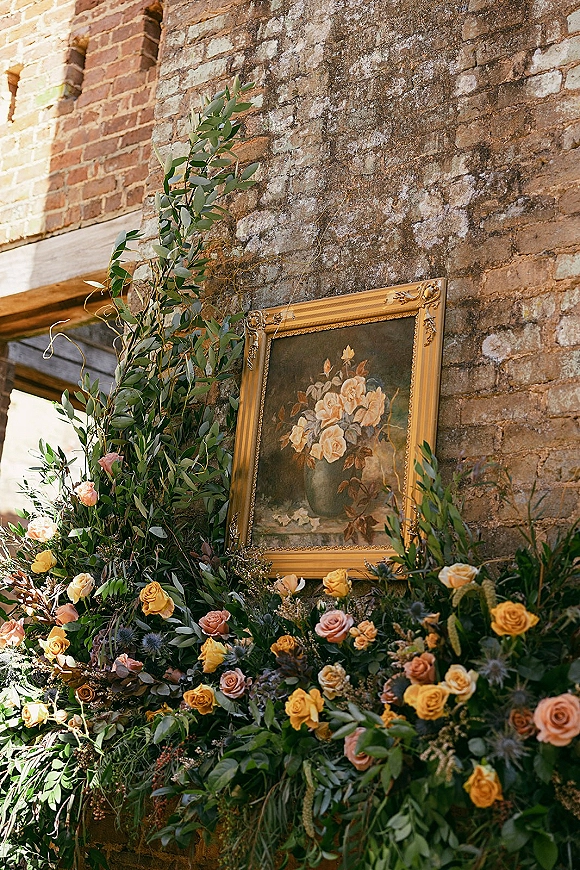 Wedding floral installation with a floral ground arrangement of roses and greenery beside a vintage gold frame against an exposed brick wall