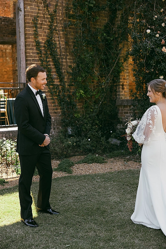 First look moment as bride holding bouquet in a long sleeve lace wedding dress faces groom in black tux by ivy brick wall in sunlight
