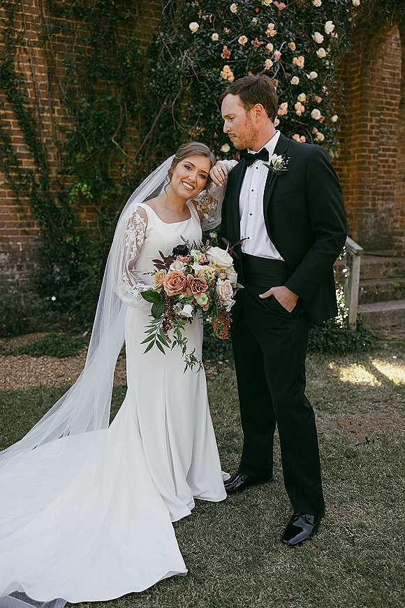 Couple portrait with bride and groom, bride leaning on groom holding a bouquet, bridal veil and lace sleeves by a rose-covered brick wall