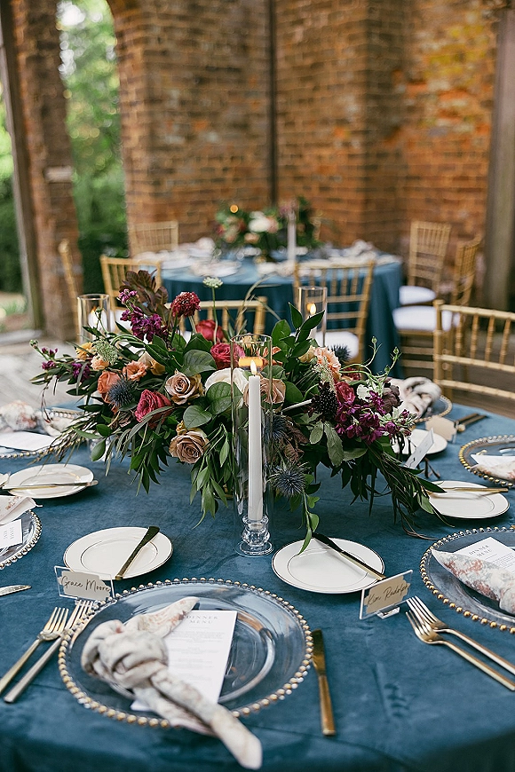 Reception tablescape with a wedding table centerpiece, taper candles in glass hurricanes, gold flatware, and blue velvet tablecloth by brick arches