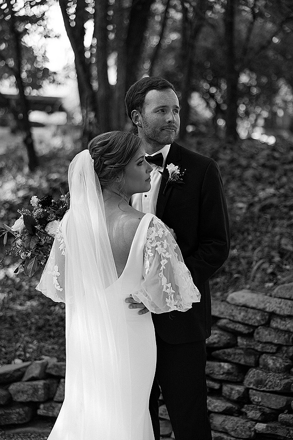 Couple portrait in a black and white wedding portrait, bride and groom embracing with veil and bouquet beside a stone wall in garden