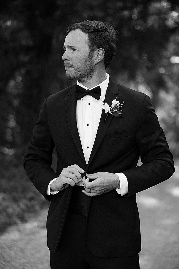 Groom portrait in tuxedo, groom in tuxedo looking away in a black tuxedo with bow tie and boutonniere on a tree-lined path