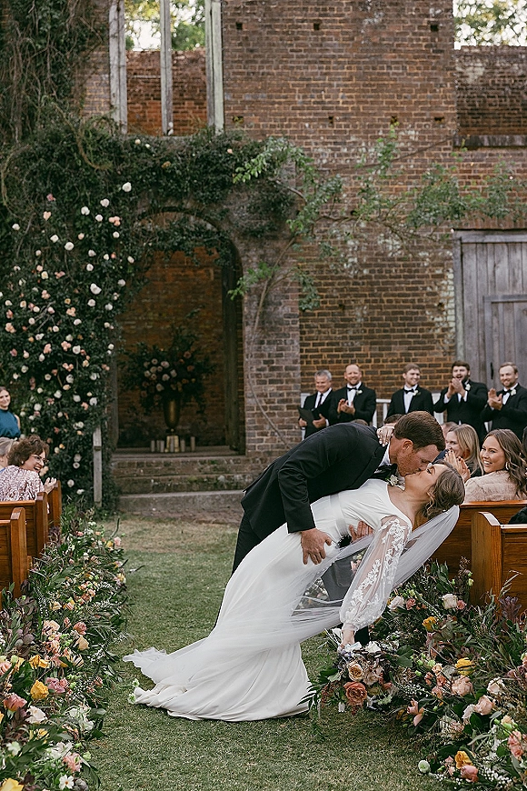 Wedding kiss as groom dips bride in veil and gown, bouquet in hand, along floral aisle before brick wall and arched doorway backdrop