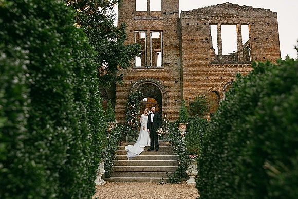 Couple portrait of bride and groom on stone steps, her veil and long train flowing beside a floral arch with greenery at brick ruins