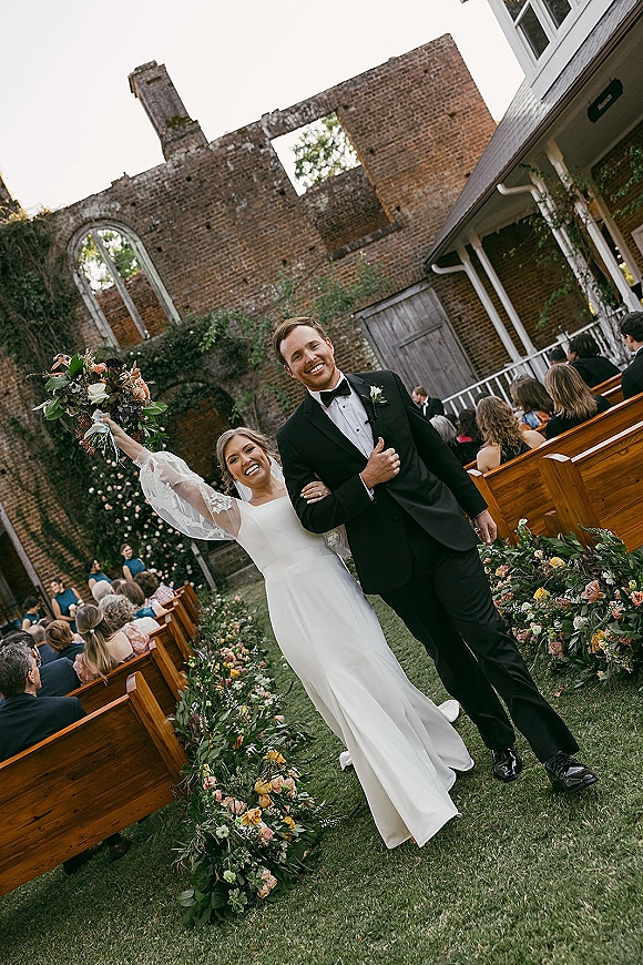 Wedding recessional as bride and groom walk the grass aisle, bride lifting bouquet and veil flowing past wooden pews and brick ruins backdrop