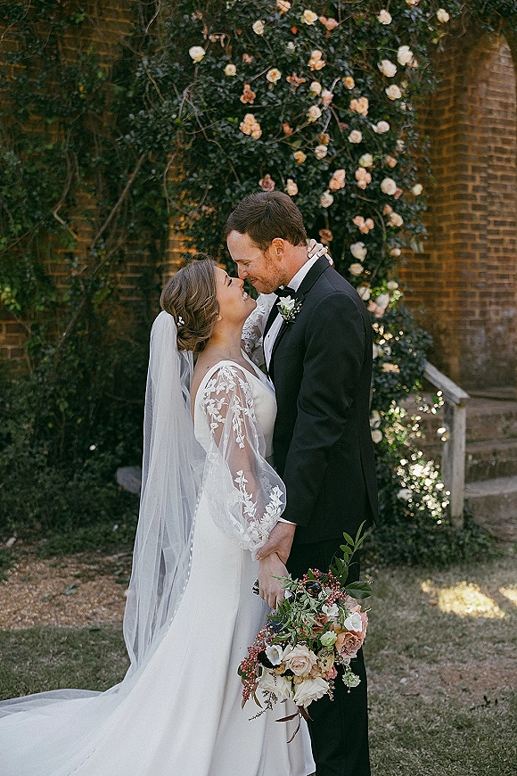 Wedding couple portrait of bride and groom forehead touch, holding hands by rose vines on a brick wall, her lace sleeves and veil flowing