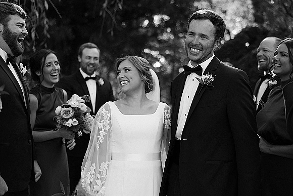 Wedding party portrait in a black and white wedding photo, bride in veil with groom and friends laughing outdoors amid soft bokeh trees