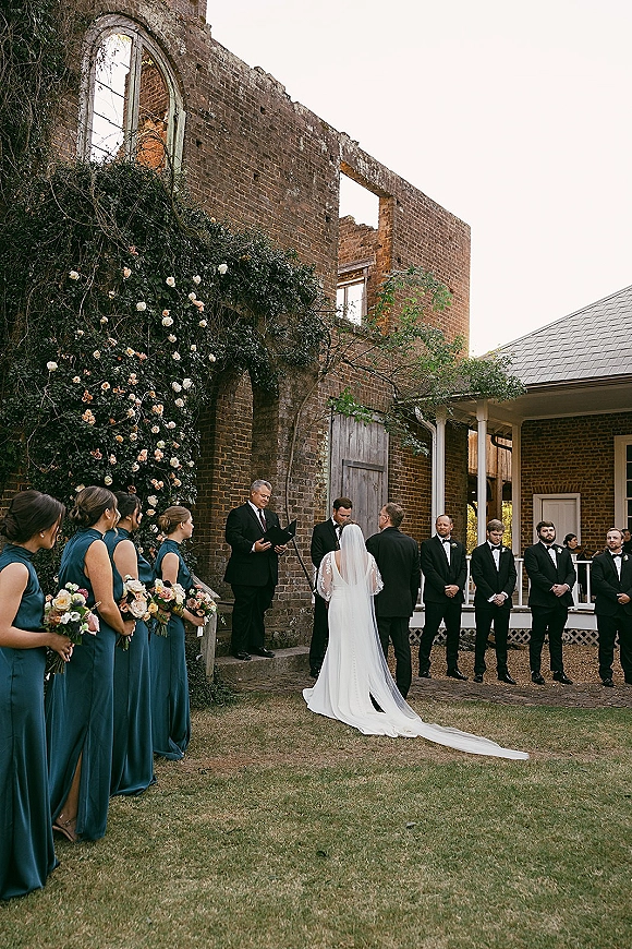 Wedding ceremony with bride and groom at the altar, bride’s long veil trailing as the party lines up before a rose-covered brick wall
