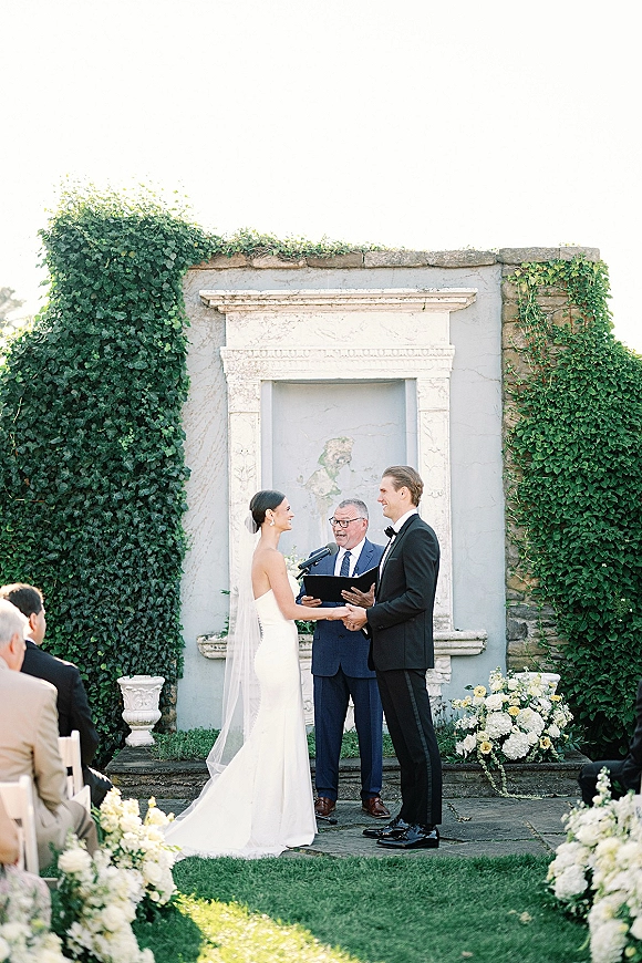 Wedding vows as bride and groom hold hands while the officiant reads from a book beside a microphone, with ivy wall backdrop
