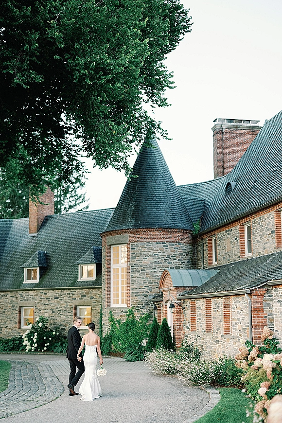 Couple portrait of bride and groom holding hands, walking away on a cobblestone driveway by a stone manor, bouquet in hand