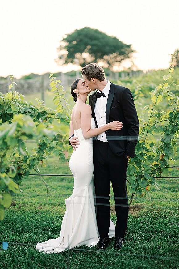 Wedding kiss portrait of bride and groom kissing in vineyard rows, groom in tuxedo with bow tie, bride in strapless dress under open sky