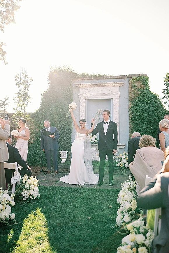 Ceremony recessional with bride and groom raising hands, bride holding bouquet up as they walk past aisle flowers in sunny courtyard
