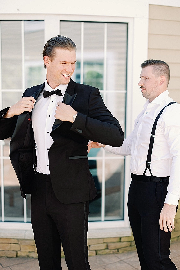 Groom getting ready in a classic black tuxedo with bow tie and suspenders, checking his wristwatch by a large window by the patio