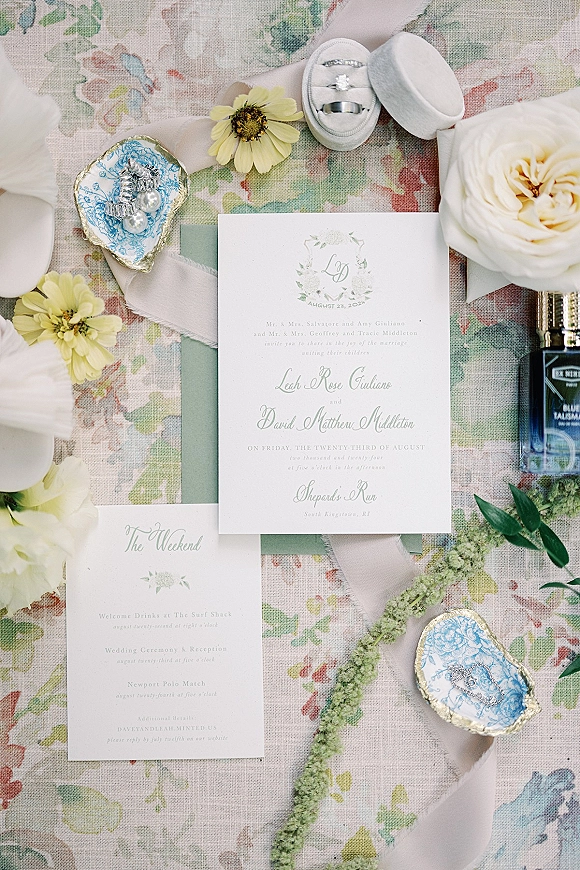 Invitation suite wedding invitation flat lay with ribbon-wrapped cards, rings in oyster shells, pearl earrings, blooms on floral linen backdrop