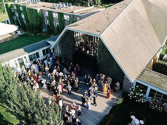 Wedding cocktail hour on an outdoor deck with guests mingling around high-top tables, bar setup, and string lights by an ivy-covered venue