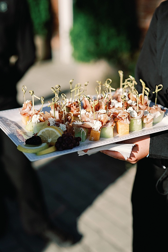 Wedding cocktail appetizers on a passed hors doeuvres platter with prosciutto, whipped cheese, fruit skewers, and lemon slices outdoors by greenery