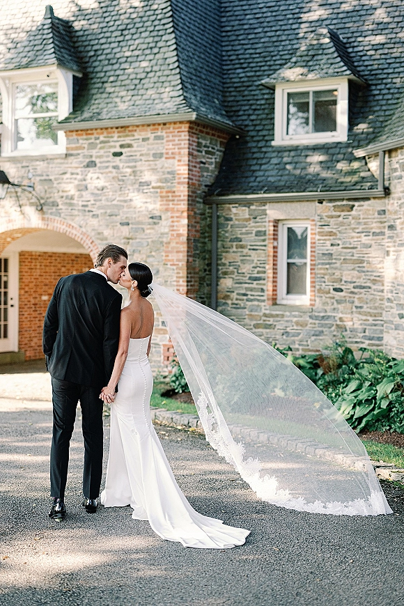 Wedding kiss portrait of the bride and groom kiss from behind, her long lace veil and train flowing on a driveway by a stone house