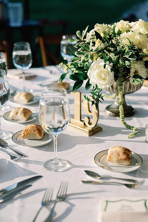 Reception tablescape with a wedding table centerpiece of white roses and greenery in a silver compote, set with gold-rim plates and water goblets