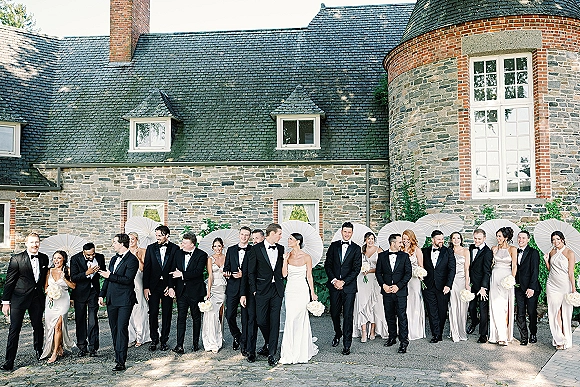 Wedding party portrait with bride and groom holding bouquets under white parasols, lined up on a cobblestone drive by an ivy-covered stone building