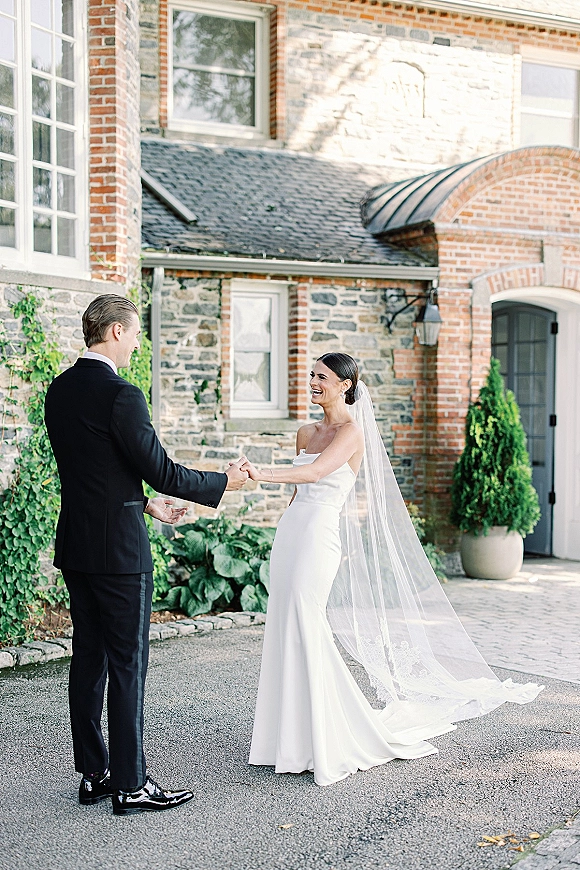 First look moment as bride in strapless wedding dress and long veil holds groom’s hands outside a stone manor with arched doorway greenery