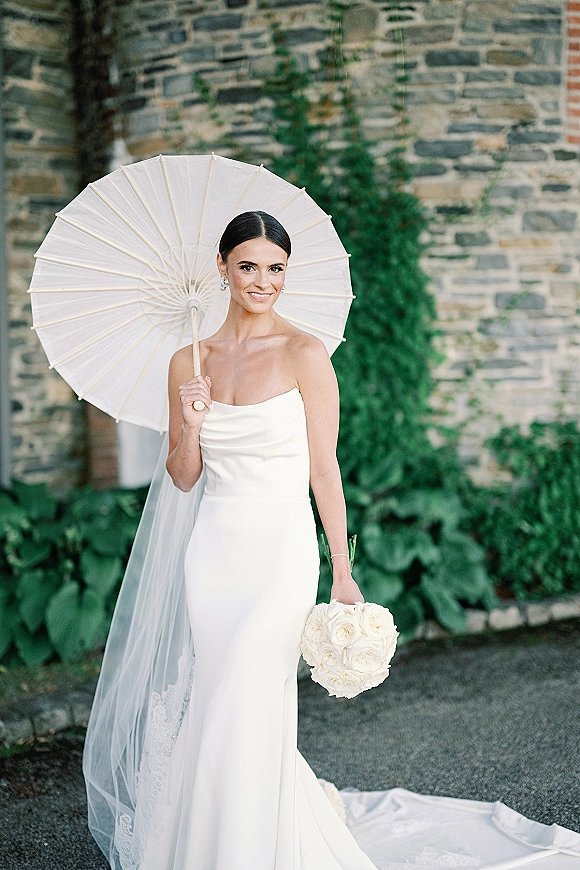 Bridal portrait of a bride with parasol holding a white rose bouquet, wearing a strapless gown and cathedral veil by an ivy stone wall outdoors