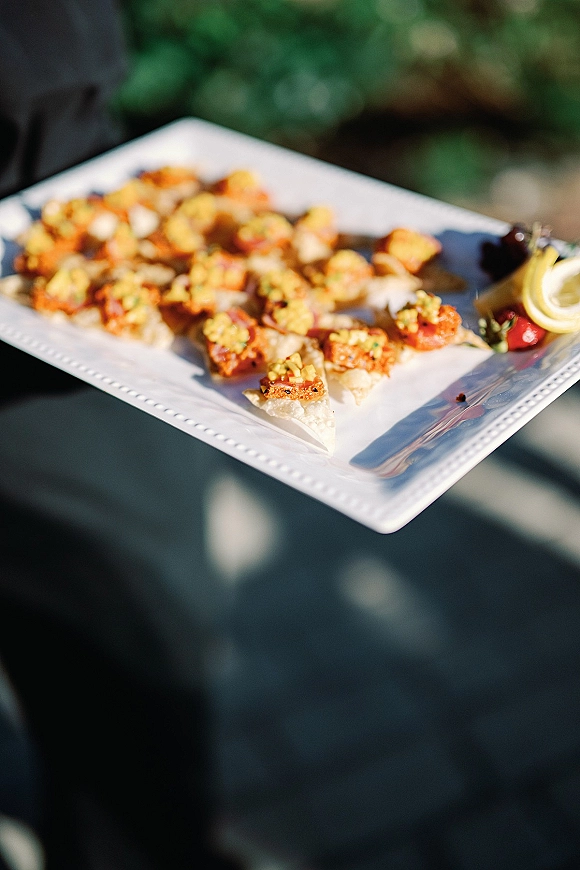 Wedding appetizers on a white square plate with bite-size shrimp, strawberries, and a lemon wedge, held during outdoor cocktail hour with guests blurred behind