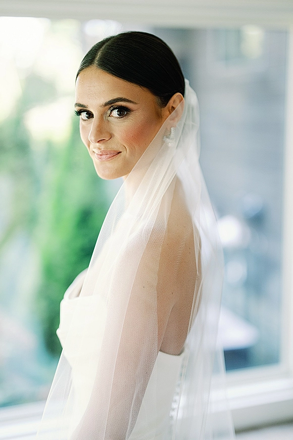 Bridal portrait of a bride looking over her shoulder in a strapless wedding dress with veil and stud earrings in soft window light