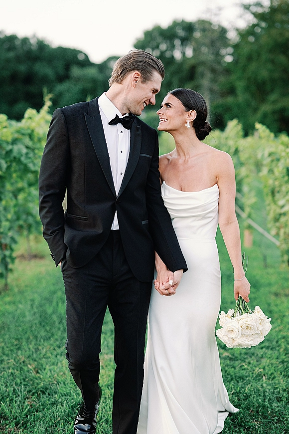 Couple portrait of bride and groom holding hands, bride in strapless gown with white rose bouquet walking through a vineyard lawn