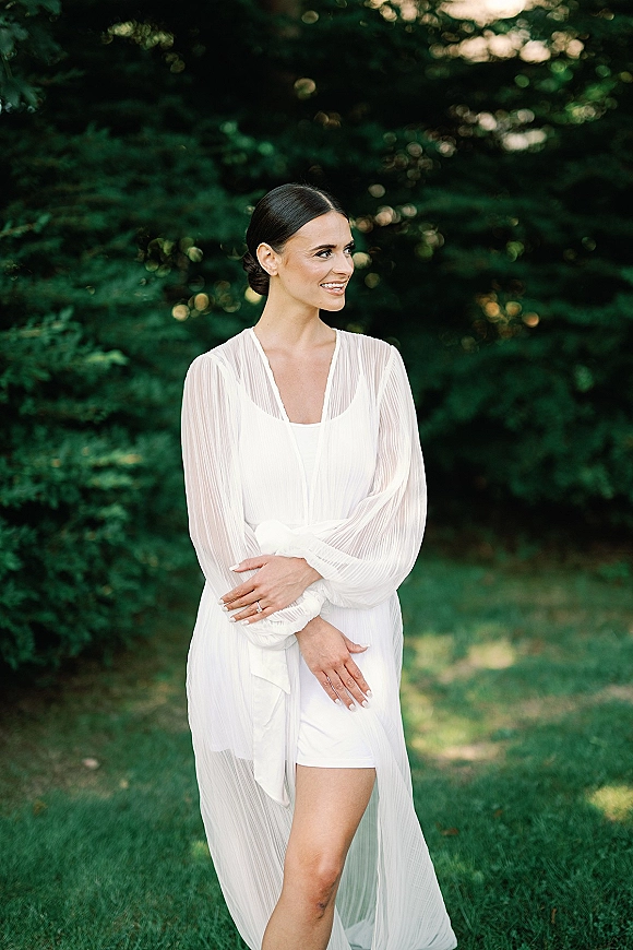 Bridal portrait of a bride in a white V-neck gown with long sheer sleeves, hands crossed showing an engagement ring in a garden setting