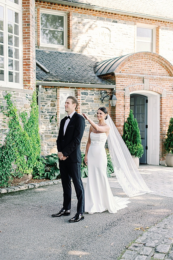Wedding first look as bride taps groom’s shoulder, her long veil and strapless dress train flowing in an ivy courtyard by an arched doorway