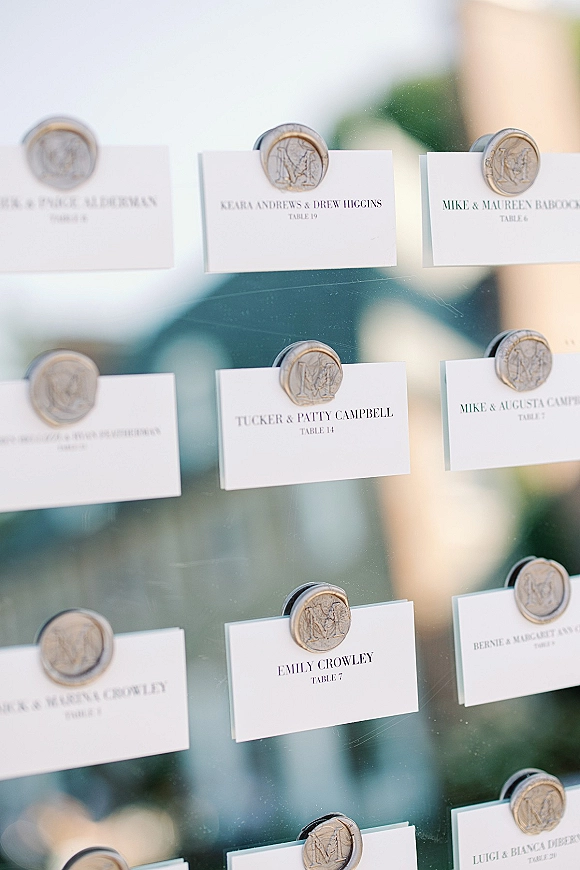 Wedding seating chart with escort card display featuring white place cards and monogram wax seals on acrylic board, greenery behind
