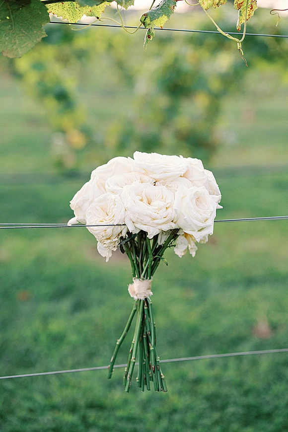 Bridal bouquet of white roses with ribbon wrap hanging on a wire fence in a vineyard, stems visible against soft green grass background