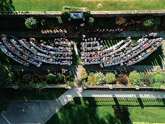 Outdoor wedding ceremony with curved white chairs and an aisle runner, framed by floral arrangements and greenery on a garden lawn by a stone wall
