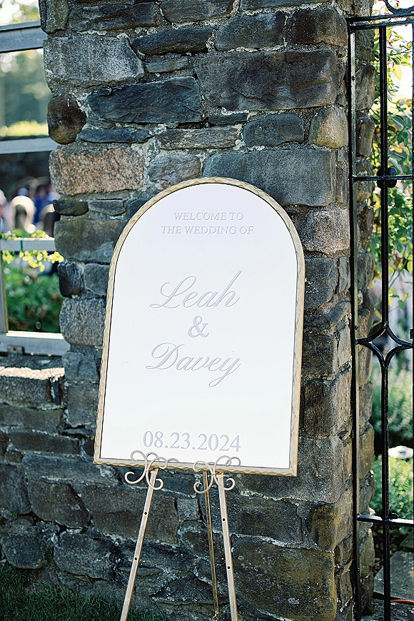 Wedding welcome sign with an arched gold frame and calligraphy lettering on an easel, set by a stone wall and wrought iron gate outdoors