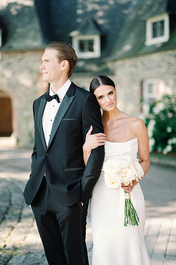 Couple portrait of a bride holding groom’s arm with a white rose bouquet, strapless dress and black tuxedo on a stone estate driveway