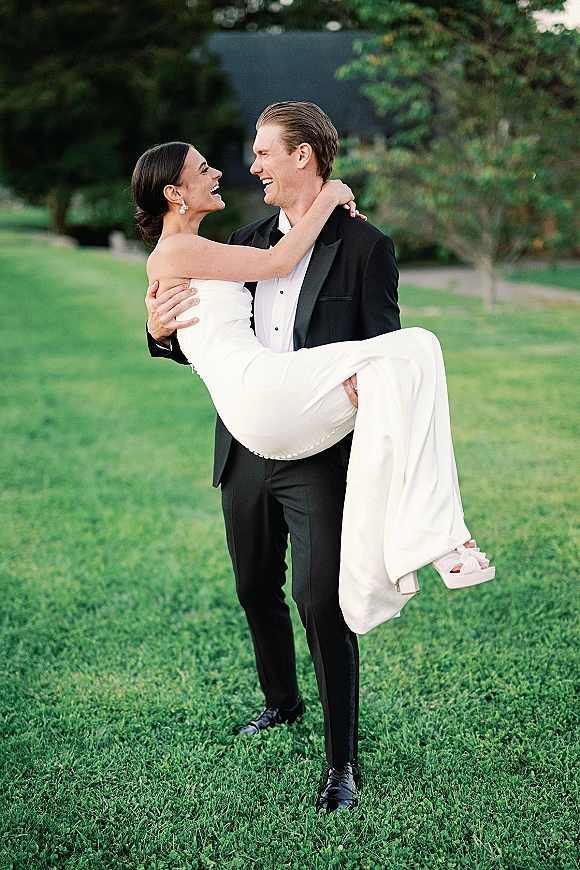 Couple portrait of groom carrying bride in his arms, both laughing, her strapless dress and drop earrings on a garden lawn with trees