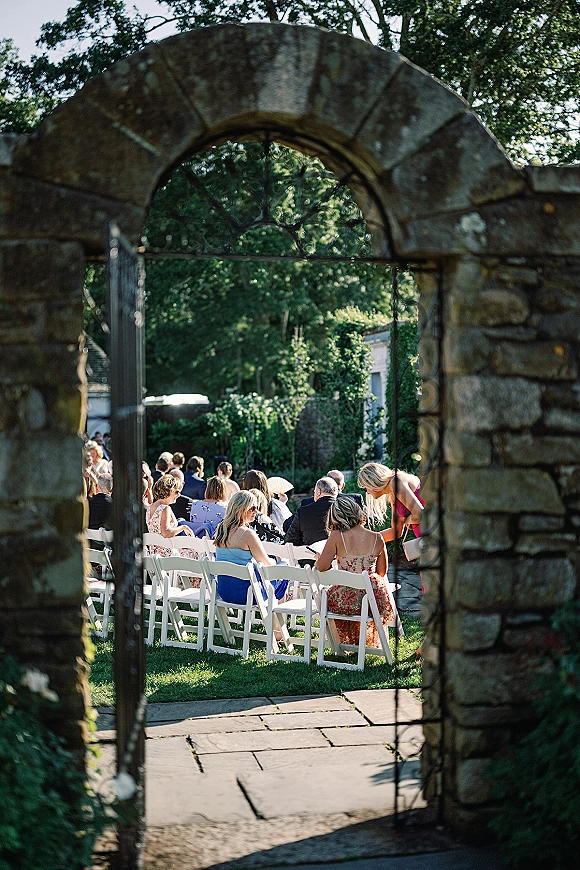 Outdoor ceremony seating with white folding chairs and wedding guests facing a stone archway and wrought iron gate on a garden lawn