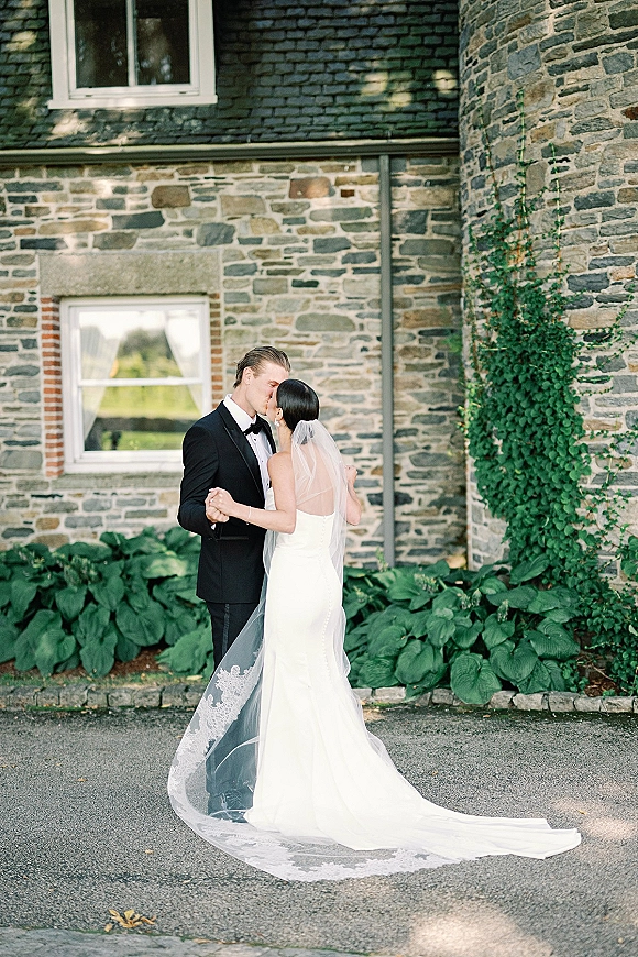 Wedding kiss portrait of bride and groom kiss, holding hands in tuxedo and strapless gown with long lace veil train by an ivy stone wall
