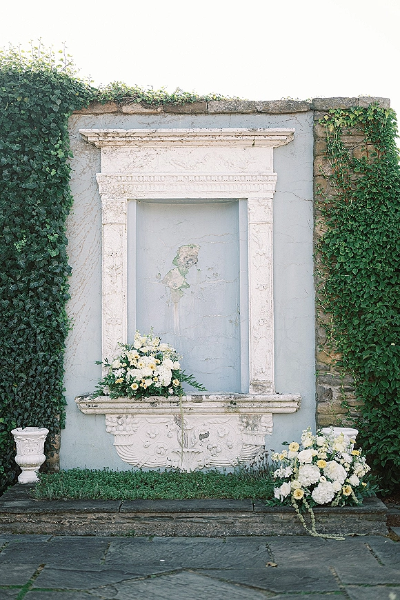 Wedding ceremony backdrop with white and yellow floral arrangements and greenery garland on a carved stone niche wall with ivy vines